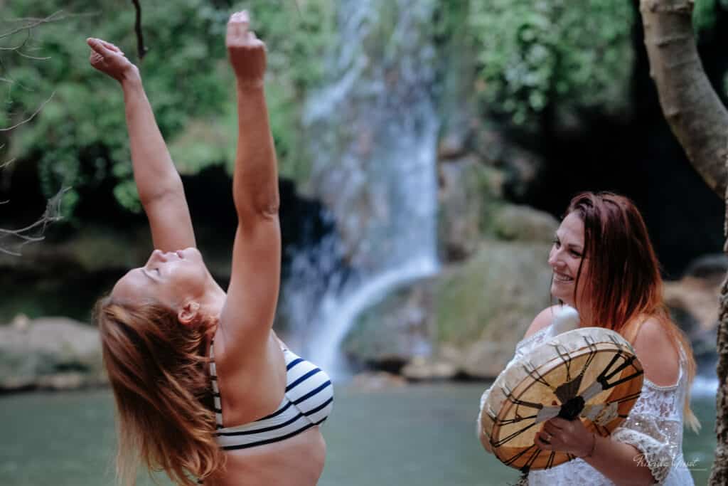 femme qui joue au tambour devant une femme qui danse les yeux fermé les bras levé au ciel devant une cascade