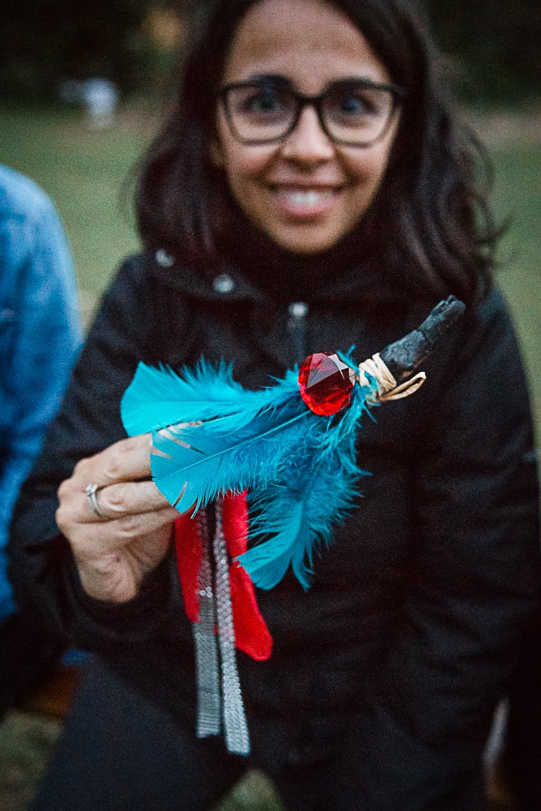 femme avec des lunettes qui sourie en montrant un bâton décoré de plume bleu et pierre rouge