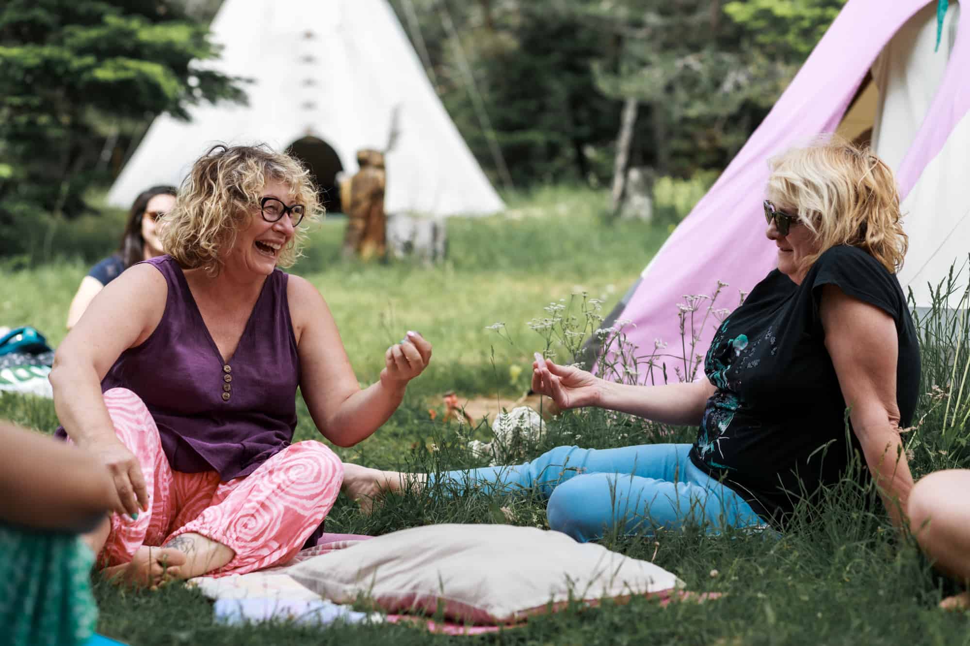deux femmes assises dans l'herbe devant un Tipi qui rigolent