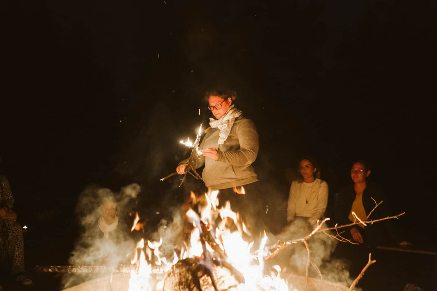 femmes devant un feu de camp avec une qui tien un bout de bois en feu