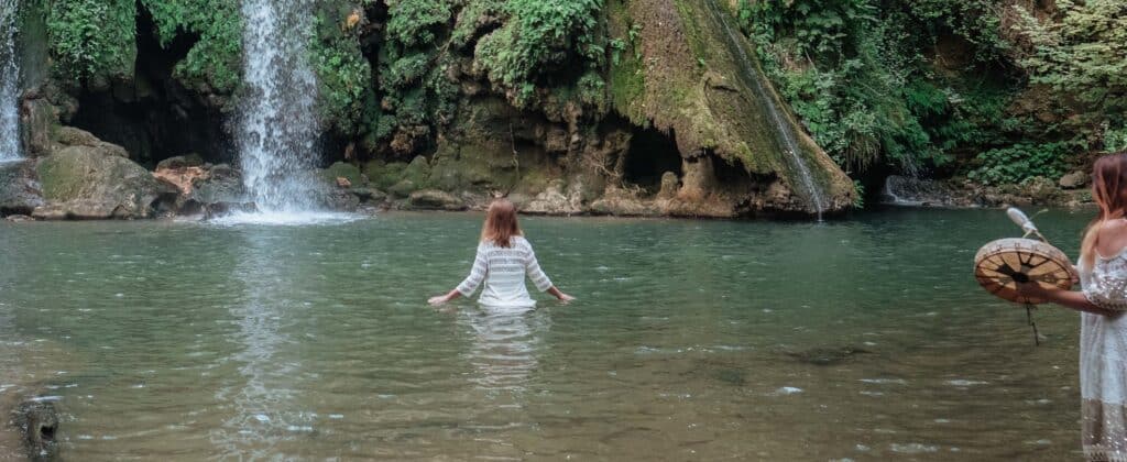 femme qui avance en robe blanche dans le bassin d'une cascade et une autre qui joue au tambour