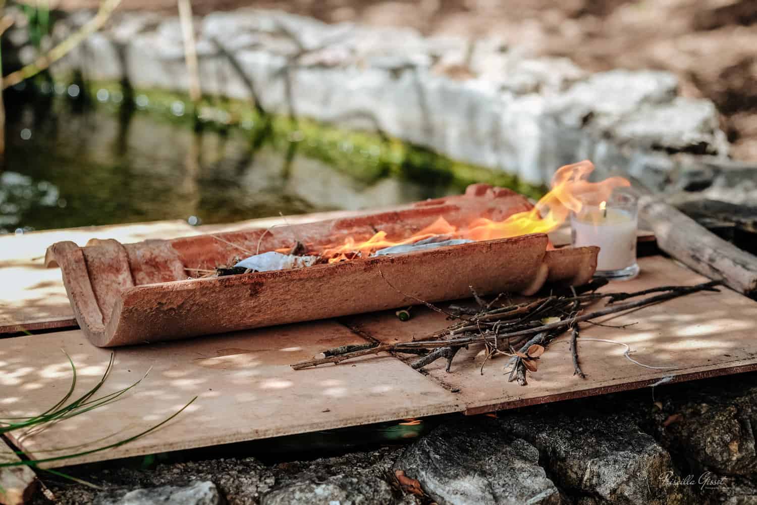 du feu dans une tuile posée sur un murée au bord de l'eau avec une bougie a coté et des brindilles des bois