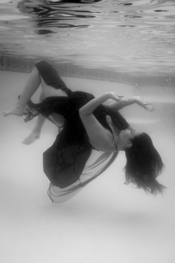 Femme en mouvement sous l’eau, vêtue d’une robe noire flottante, lors d’une séance de photo-thérapie.